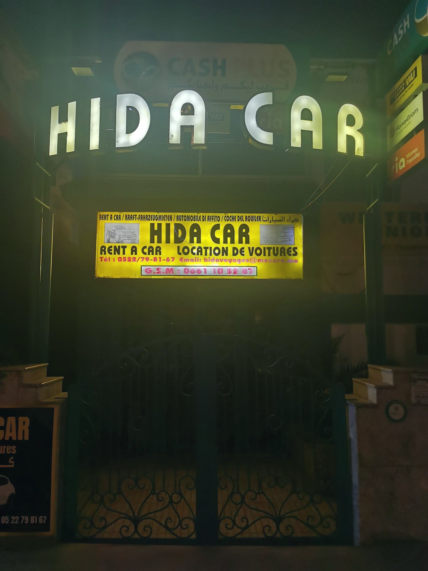 La devanture de nuit : lettres HIDA CAR allumées au-dessus de l’enseigne jaune, grille fermée